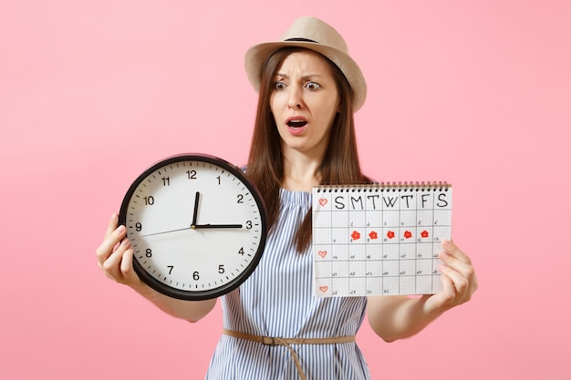 Shocked confused sad woman in blue dress holding round clock, periods calendar for checking menstruation days isolated on trending pink background. Medical healthcare gynecological concept. Copy space