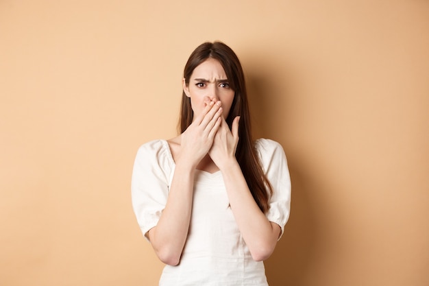 Shocked and confused woman frowning upset, covering mouth with hands, disappointed from something terrible, standing on beige background.