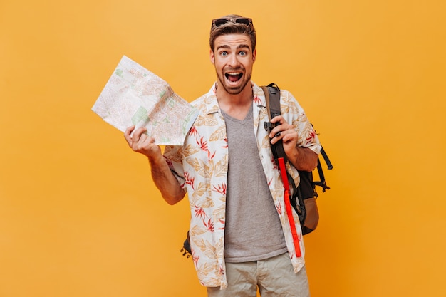 Shocked cool guy with modern hairstyle and red beard in painted summer outfit holding map and screaming on orange wall