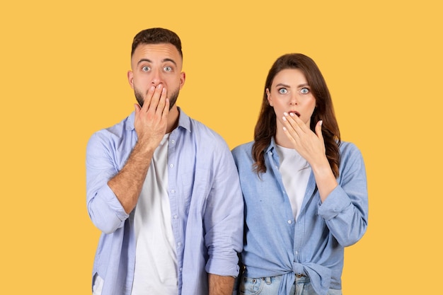 Shocked couple covering mouths wideeyed surprise on yellow background