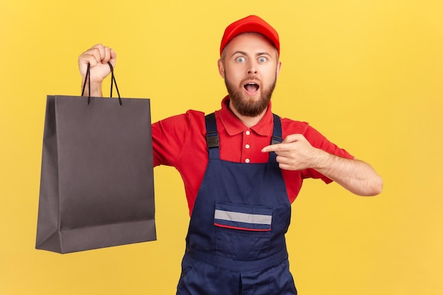 Shocked courier man wearing blue uniform pointing at black shopping bag delivering order to client