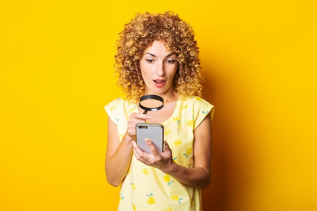 Giovane donna riccia scioccata che guarda con una lente d'ingrandimento nel telefono su uno sfondo giallo