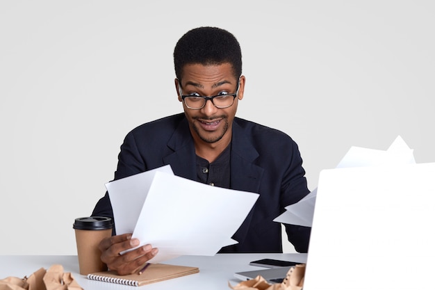 Shocked dark skinned man carries many papers, looks surprisingly through spectacles, wears formal clothes and spectacles, sits at dektop, isolated on white. People and work concept