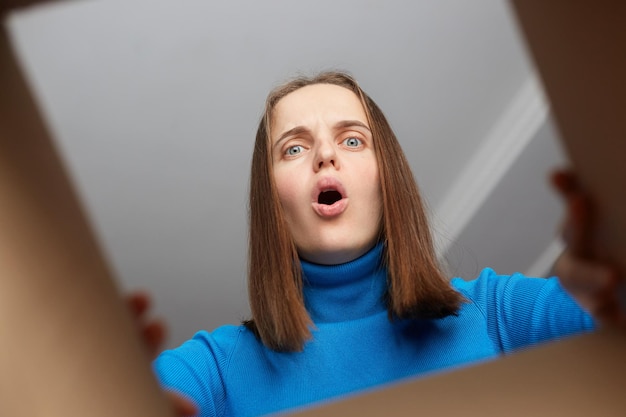 Shocked disappointed woman customer wearing blue turtleneck opening cardboard box with broken or wrong order looking inside parcel with widely open mouth and big eyes