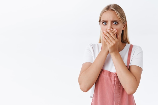 Shocked and disgusted young uneasy blond girl in pink overalls tshirt gasping cover mouth with palm from fear and panic stare camera upset standing distressed and troubled white background