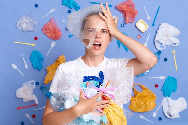 Shocked displeased woman wearing white shirt isolated on blue wall with garbage around frowning face being unhappy clean up trash astonished from pollution