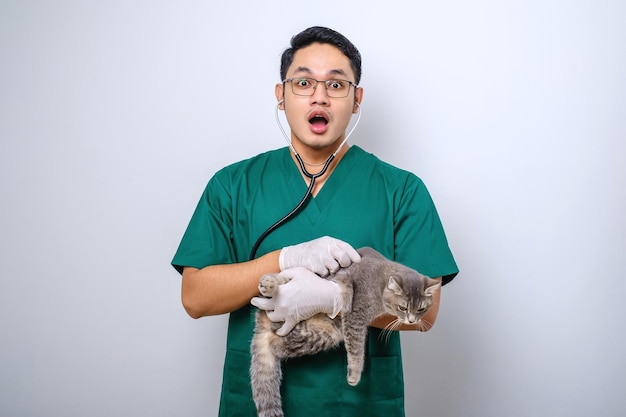 Shocked doctor in vet clinic examining cat with stethoscope gasping amazed while holding cat in his arm