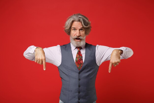 Shocked elderly gray-haired mustache bearded man in classic shirt vest and colorful tie isolated on red background in studio. People lifestyle concept. Mock up copy space. Pointing index fingers down.