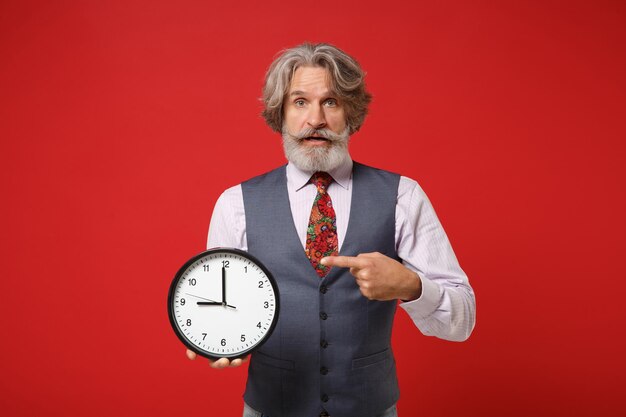 Shocked elderly gray-haired mustache bearded man in classic shirt vest and tie isolated on red wall background in studio. People lifestyle concept. Mock up copy space. Pointing index finger on clock.