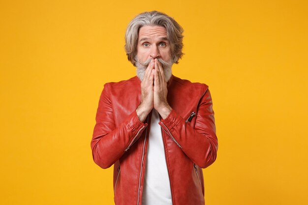 Shocked elderly gray-haired mustache bearded man in red leather jacket posing isolated on yellow orange background in studio. people lifestyle concept. mock up copy space. covering mouth with hands