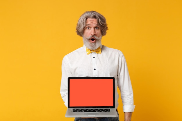 Shocked elderly gray-haired mustache bearded man in white shirt\
bow tie isolated on yellow background. people lifestyle concept.\
mock up copy space. holding laptop pc computer with blank empty\
screen.