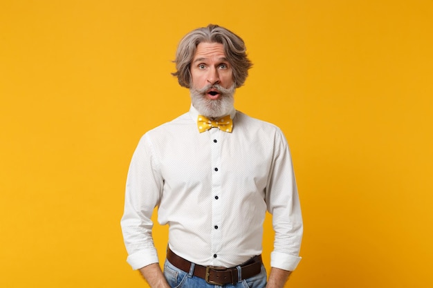 Shocked elderly gray-haired mustache bearded man in white shirt bow tie posing isolated on yellow orange background, studio portrait. People lifestyle concept. Mock up copy space. Keeping mouth open.