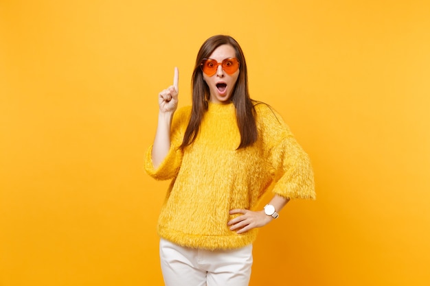 Shocked excited young woman in sweater, heart orange glasses pointing index finger up on copy space isolated on bright yellow background. people sincere emotions, lifestyle concept. advertising area