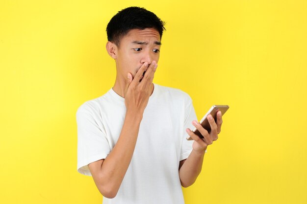 Shocked face of Asian man in white shirt looking at phone screen on yellow background.