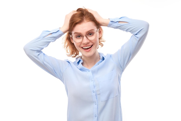 Шокированная рыжая девушка в деловой рубашке выглядит удивленной с открытым ртом, держащим голову руками на белом фоне студии