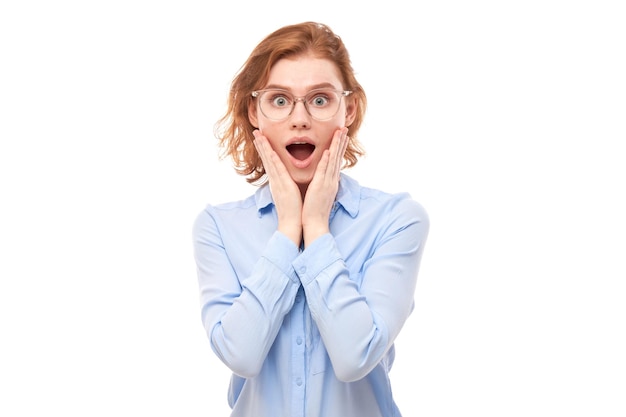 Shocked face ginger girl in business shirt looks surprised with open mouth holding head with hands on white studio background