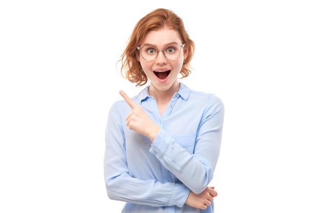 Shocked face redhaired girl in business shirt looks surprised with open mouth on white studio background points finger at copy space