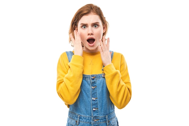 Shocked face redhaired girl looks surprised with open mouth holding head with hands on white studio background