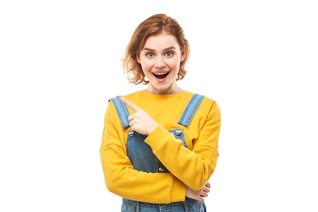 Shocked face redhaired girl looks surprised with open mouth on white studio background points finger at copy space