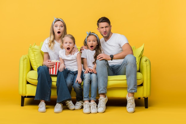 Foto famiglia scioccata che guarda film sul divano con un secchio di popcorn su giallo