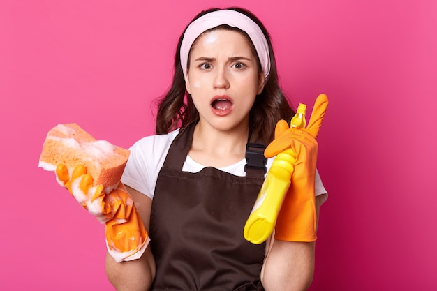 Шокированная женщина-домохозяйка держит губку и моющее средство, будучи в панике, имеет много помещений для уборки, нуждается в помощи