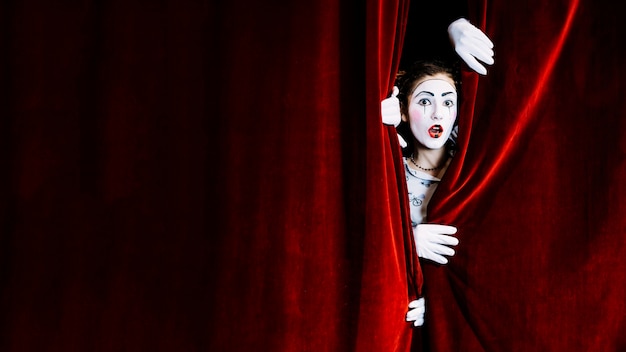 Shocked female mime artist peeking from red curtain