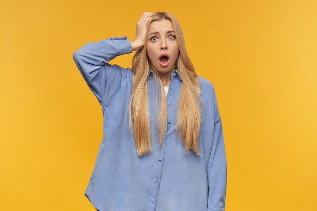 Shocked girl, stressed looking woman with blond long hair. wearing blue shirt. people and emotion concept. touching her head, forgot something. watching at the camera, isolated over orange background