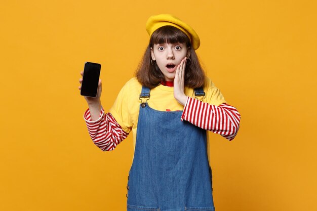 Shocked girl teenager in french beret put hand on face, holding mobile phone with blank empty screen isolated on yellow wall background. People sincere emotions, lifestyle concept. Mock up copy space.