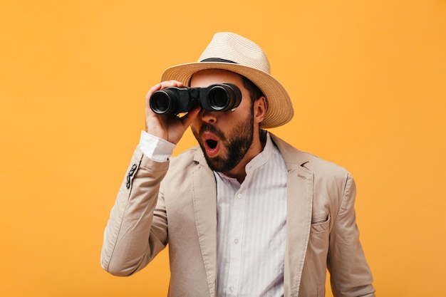 Shocked guy in hat looks into binoculars on orange background