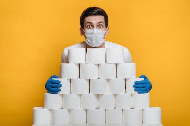 Photo shocked guy in protective mask behind a big stack of toilet paper