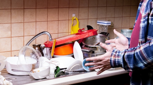 Ragazzo della mano scossa vicino a molti piatti sporchi che giacciono nel lavandino in cucina che vuoi lavare