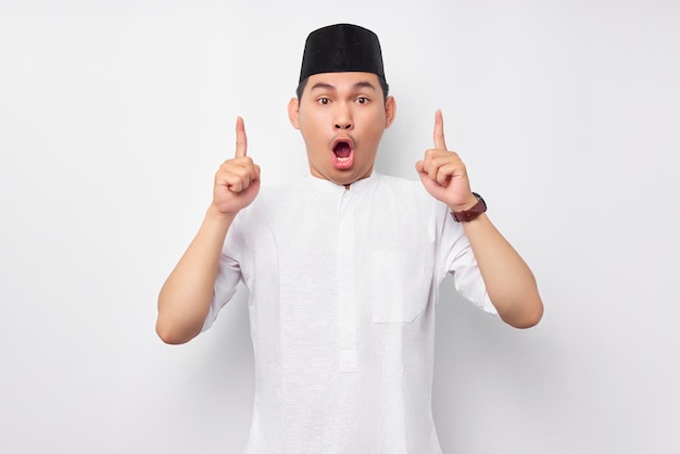 shocked handsome young asian muslim man wearing arabic costume pointing finger at free space place for an advertisement isolated on white background people religious islamic lifestyle concept