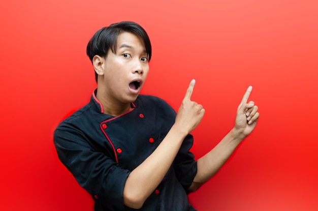 Shocked happily young asian chef in black chef uniform\
presenting something over red background.