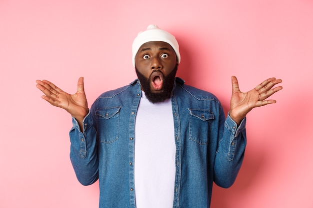 Shocked and impressed Black man staring at camera with complete disbelief, saying wow, standing in beanie and hipster shirt over pink background