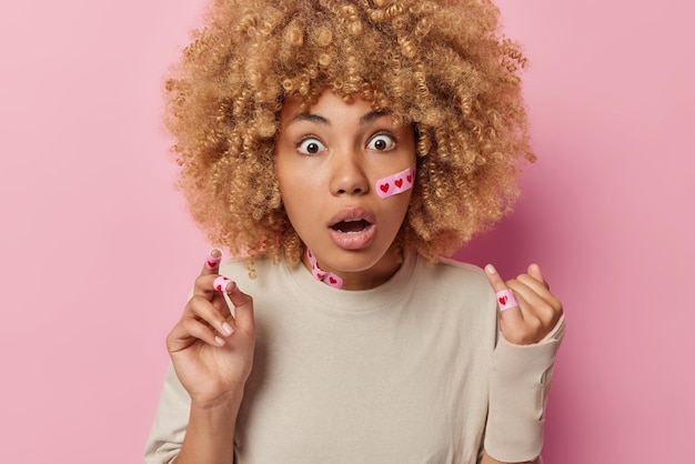 Shocked impressed young woman with curly hair puts adhesive\
plaster on finger and face got injury dressed in casual beige\
background isolated over pink background self treatment after\
trauma