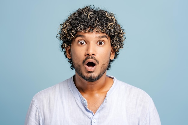 ショックを受けたインド人の若者が青い背景でポーズをとり驚きの感情を表し口を広く開けています