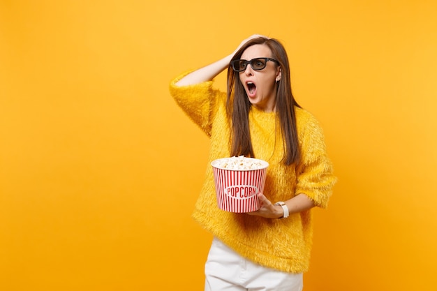 Shocked indignant young girl in 3d imax glasses putting hand on head, screaming watching movie film, hold bucket of popcorn isolated on yellow background. People sincere emotions in cinema, lifestyle.