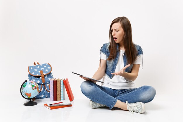 Studentessa irritata scioccata che tiene in mano un computer tablet pc, allargando la mano, seduta vicino al globo, zaino, libri scolastici isolati