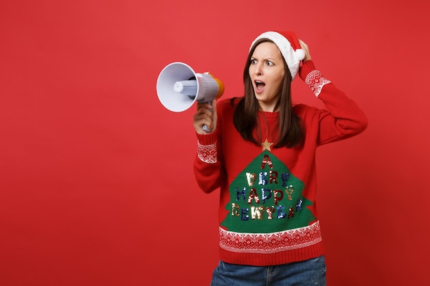 Shocked irritated young Santa girl in Christmas hat screaming on megaphone, putting hand on head isolated on red background. Happy New Year 2019 celebration holiday party concept. Mock up copy space.