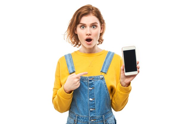 Shocked joyful redhead girl points finger at smartphone blank screen in yellow clothes isolated on white studio background