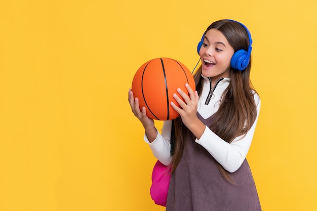 Шокированный ребенок в наушниках со школьным рюкзаком и баскетбольным мячом на желтом фоне