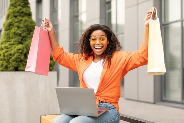 Shocked laughing young african american lady in casual and glasses with laptop picks up many packages enjoys order