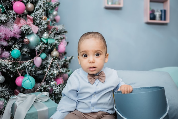 A shocked little boy in a blue shirt sits on the bed against  a Christmas tree