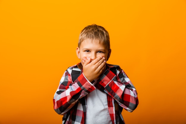 Shocked little boy in red shirt covering his mouth with his hands on yellow background