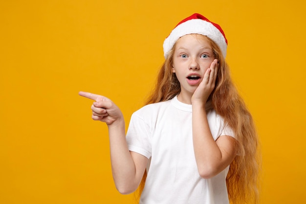 Shocked little ginger kid Santa girl 12-13 years old in white t-shirt Christmas hat isolated on yellow background. New Year 2020 celebration concept. Mock up copy space. Pointing index finger aside.