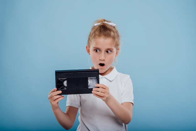 Shocked little girl with video tape isolated on blue background old gadgets