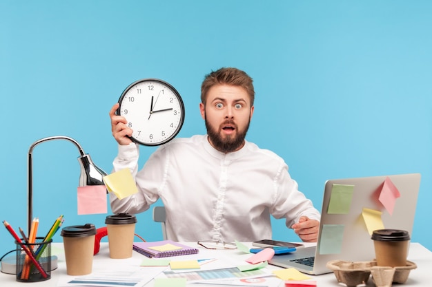 Shocked man holding wall clock and looking at camera with terrified expression sitting at workplace all covered with sticky notes, late with deadline. Indoor studio shot isolated on blue background