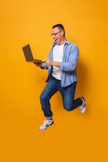 Shocked man jumping isolated over yellow wall using laptop computer.