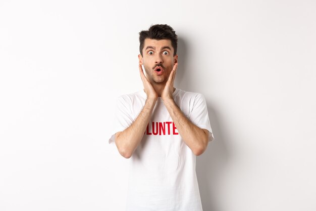 Shocked man in volunteer t-shirt gasping, staring at camera in awe, standing over white.