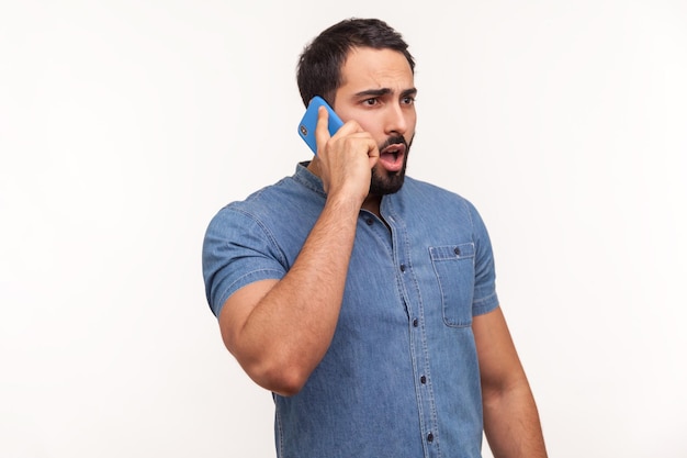 Photo shocked man with beard in blue shirt talking phone, surprised with news looking big eyes and widely opened mouth, unpleasant call, bad tariffs, roaming. indoor studio shot isolated on white background